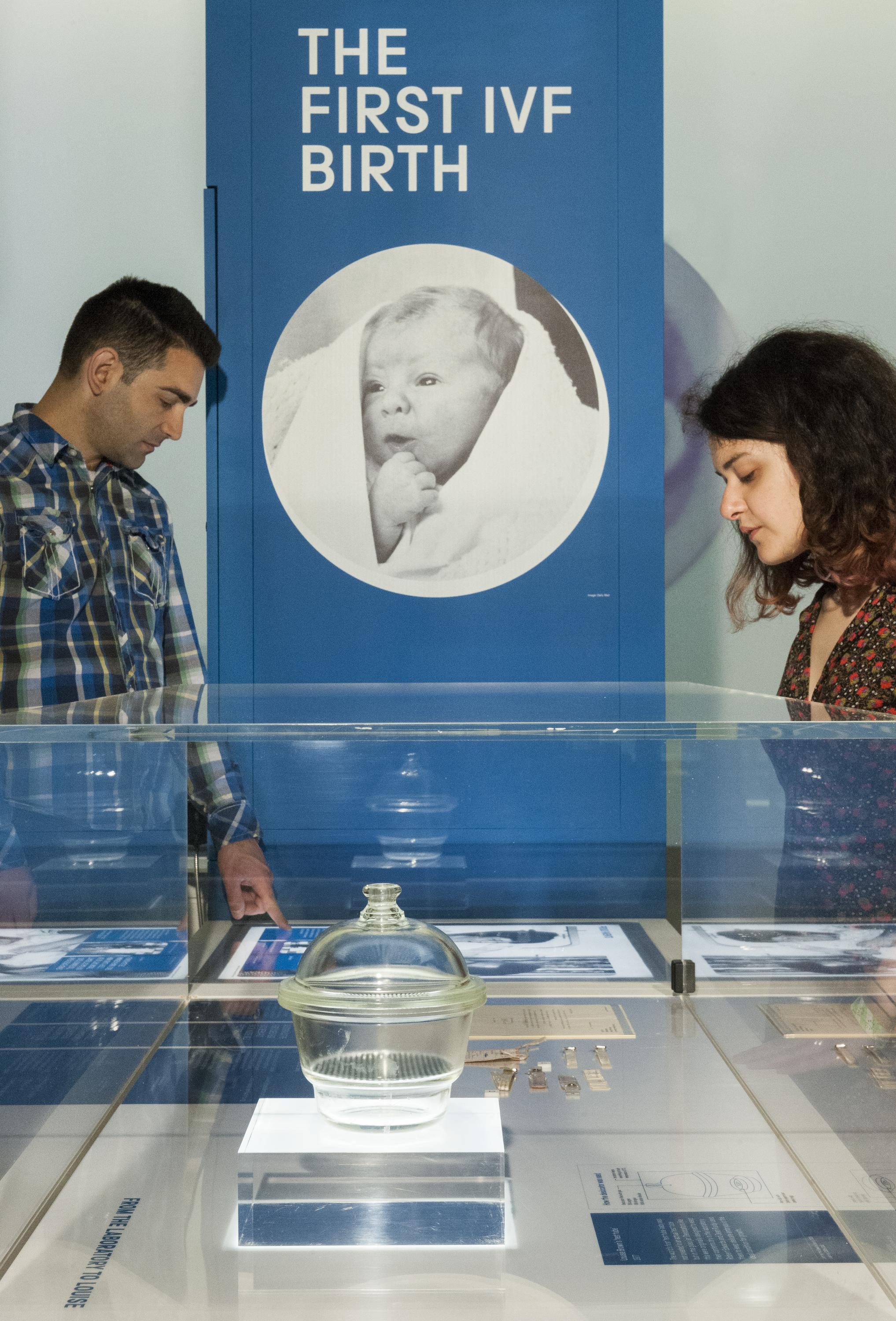 A man and a woman examine some objects in a glass case at the Science Museum's IVF exhibition. Behind them is a poster that displays an image of Louise Brown as a baby, with the text 'The first IVF birth'.